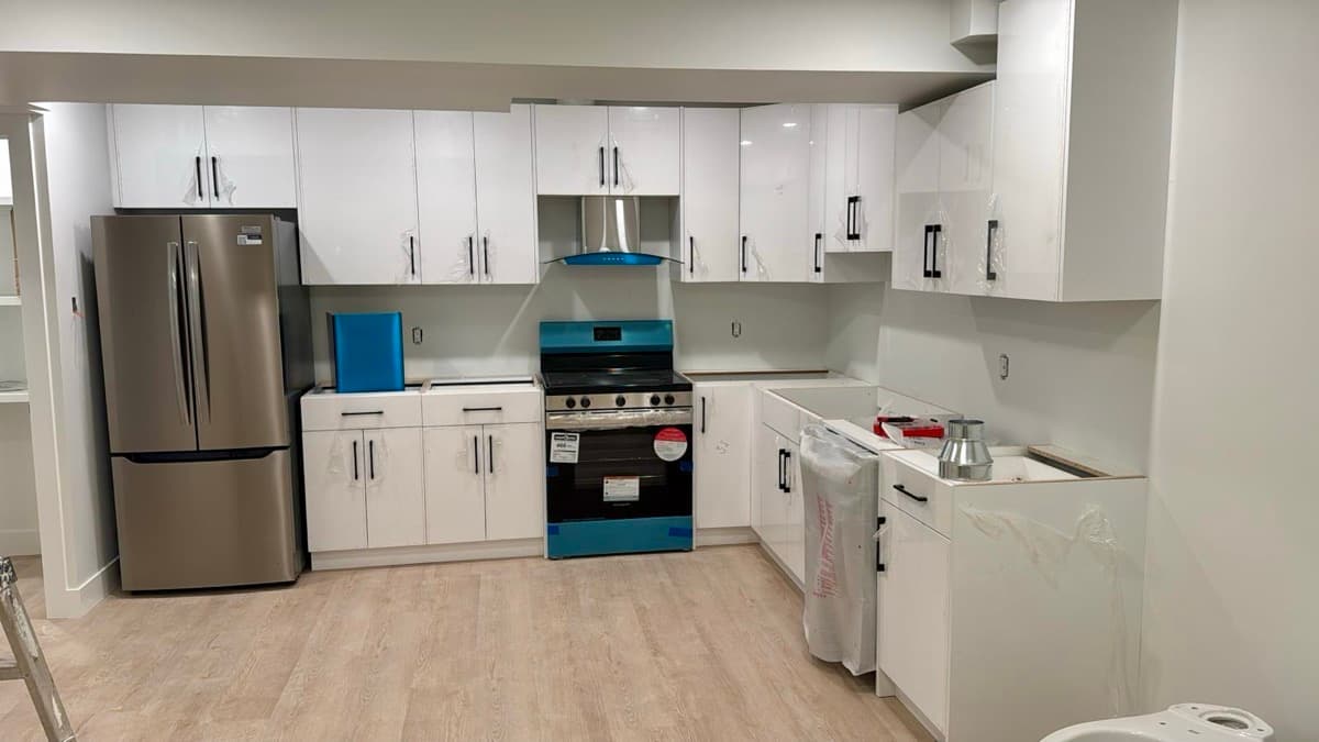 Basement kitchen during installation, with white cabinets still in protective film, a stainless fridge and range, and plank flooring