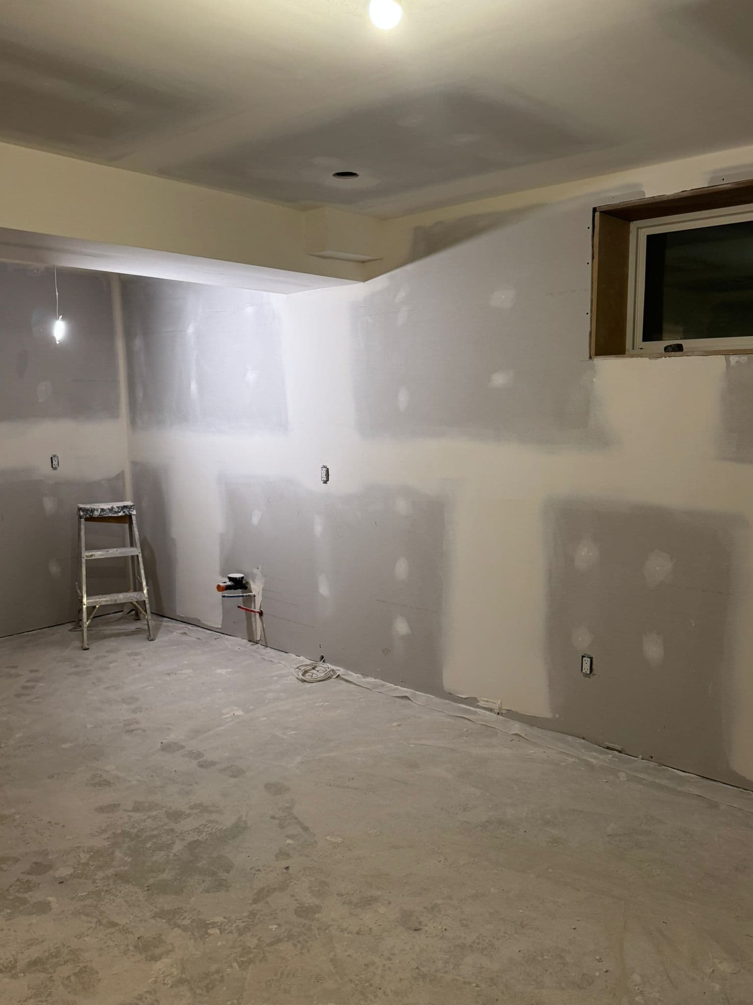 Basement living area with taped and mudded drywall, a stepladder, capped plumbing lines and a wood-framed window opening