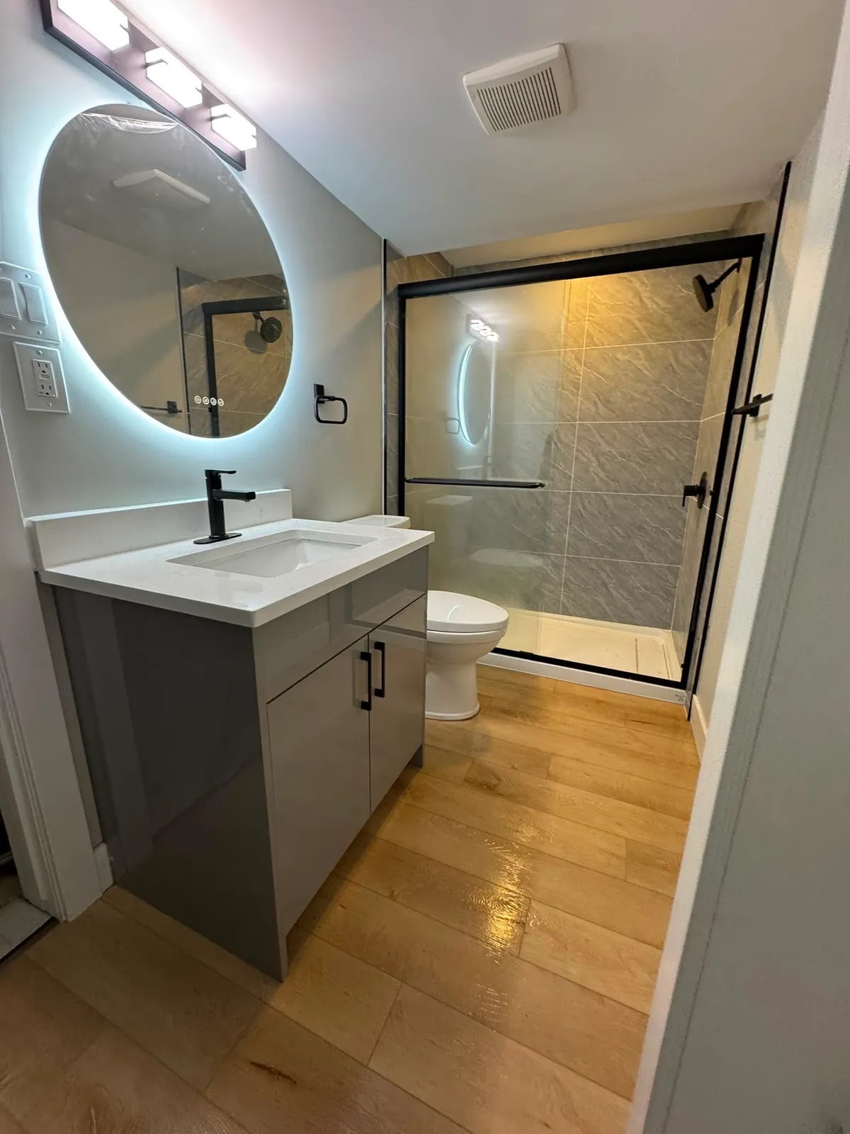Renovated bathroom with a grey gloss vanity, round backlit mirror, wood-look plank floor and a black-framed sliding shower door