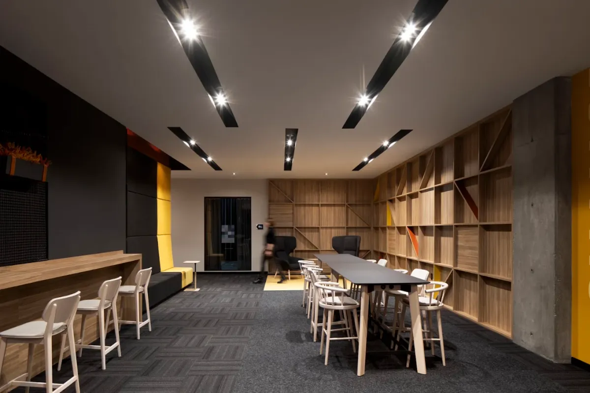 Lounge with wood shelving along one wall, a long high table with white stools, yellow accent panels and linear ceiling lights
