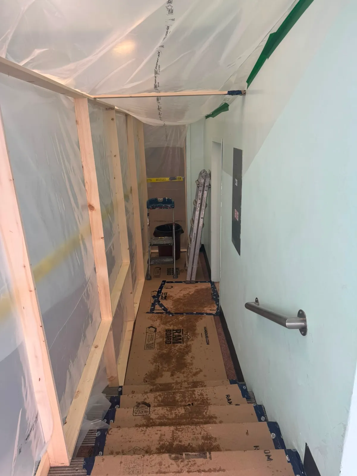 Narrow hallway and stairs covered in protective board, with plastic sheeting stapled to a wood frame down one side