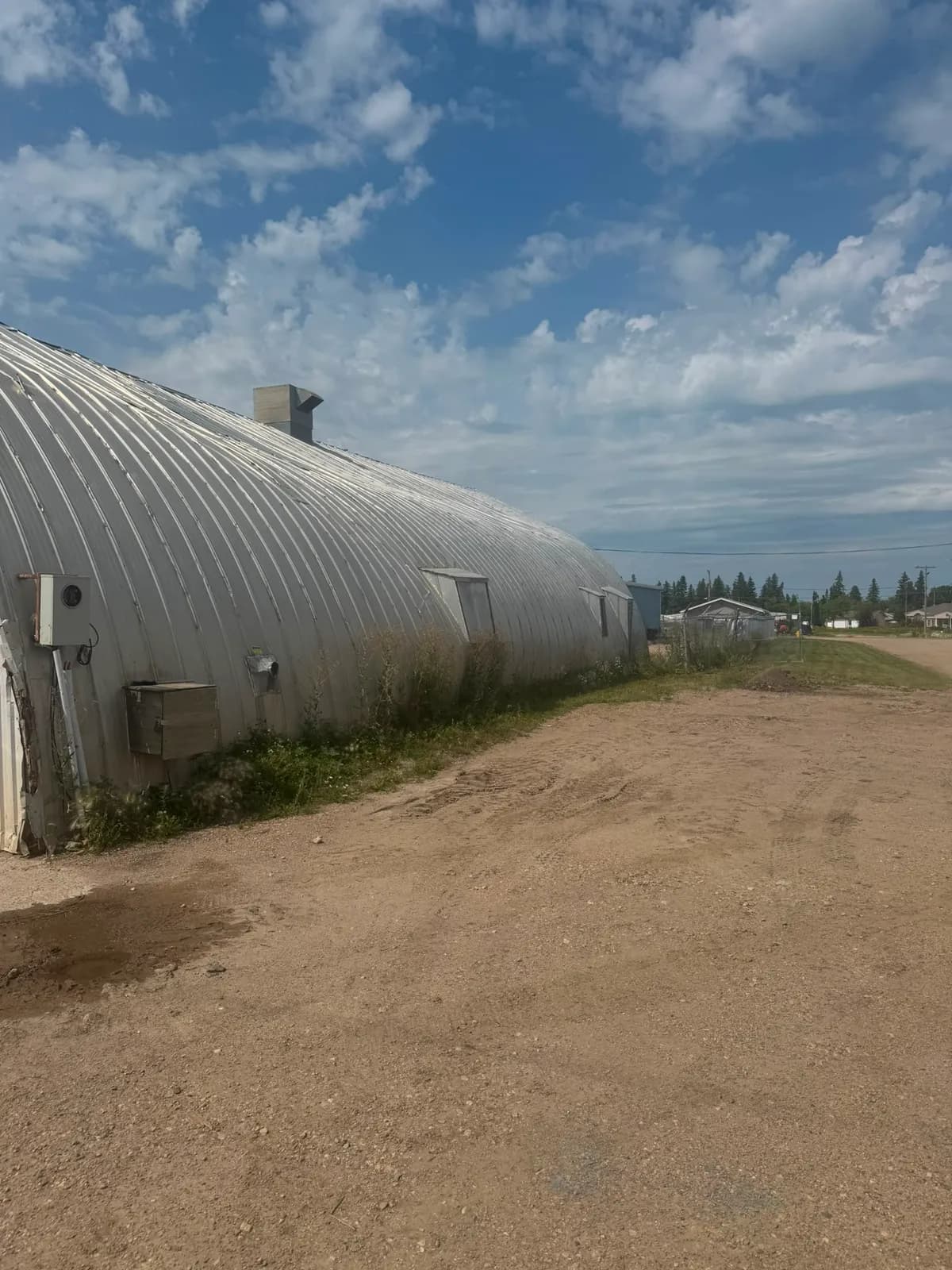Long side of an arched steel building, with an electrical meter, small vent hoods and a gravel yard alongside