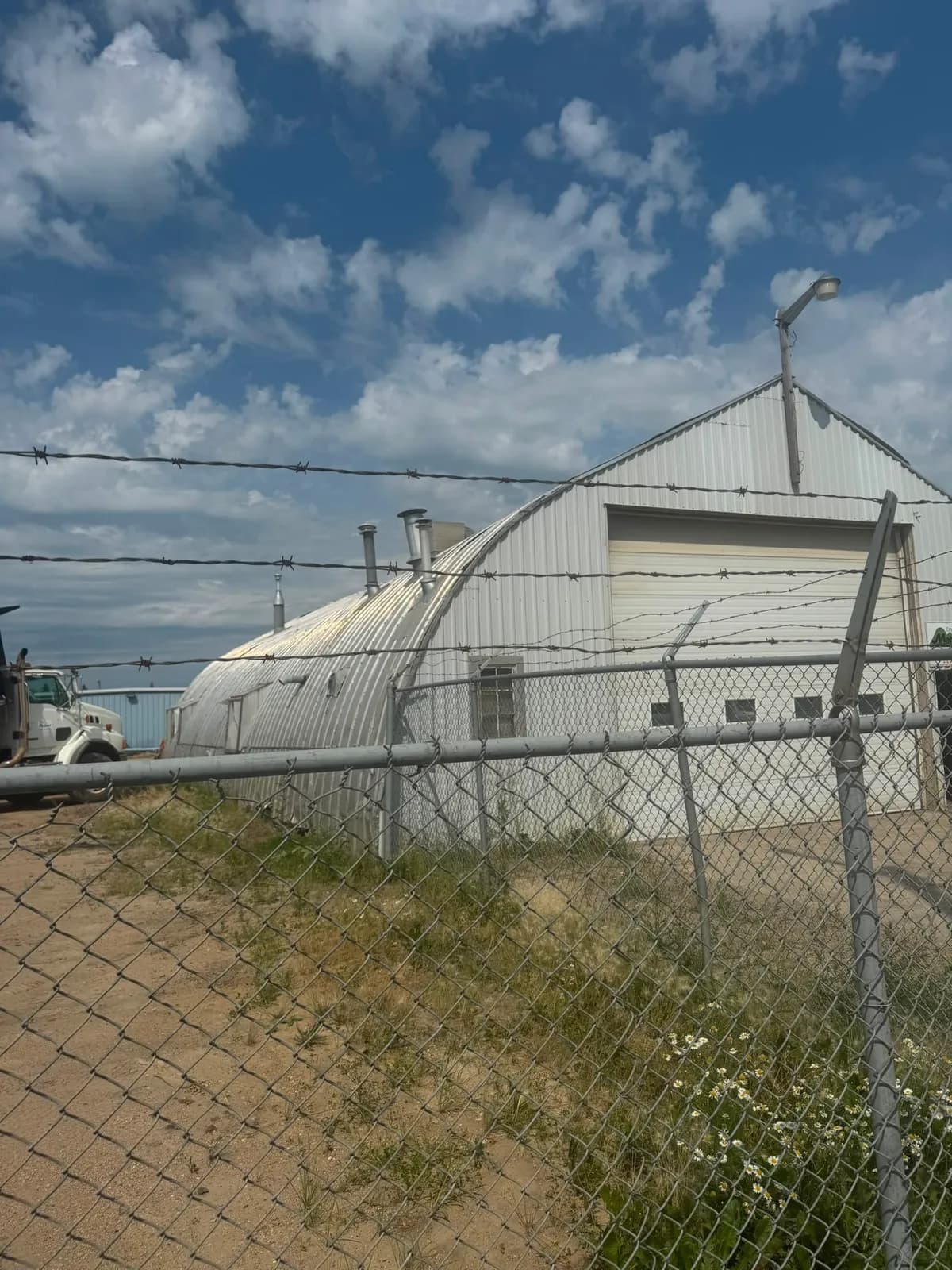 Arched corrugated steel building with an overhead door in its end wall, seen through a chain-link fence topped with barbed wire