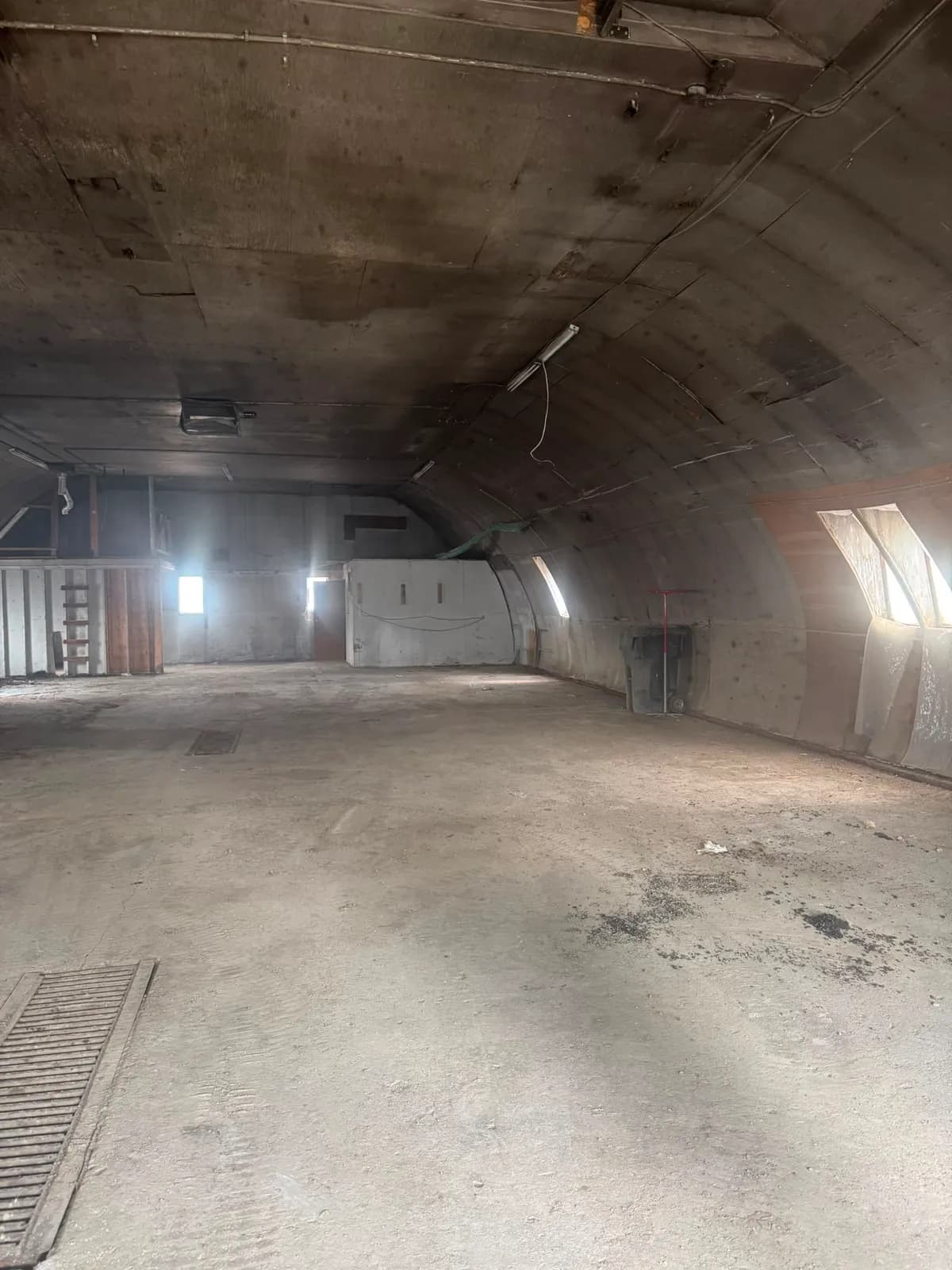 Open interior with a dusty floor slab, a floor grate, wood framing at one end and daylight from the side windows