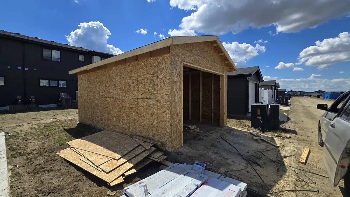 New single-car detached garage framed and sheathed in OSB with roof trusses on, beside a back lane in a new subdivision