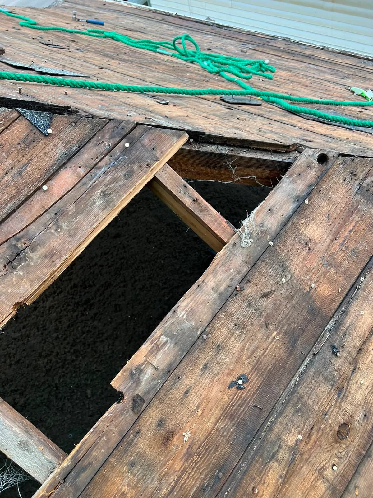 Broken roof boards pulled back to show a hole through the decking, with a green safety rope across the roof