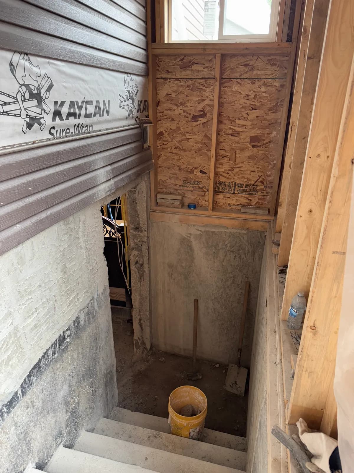 Concrete steps leading down to a new doorway cut through a basement foundation wall, with framed walls and a window above
