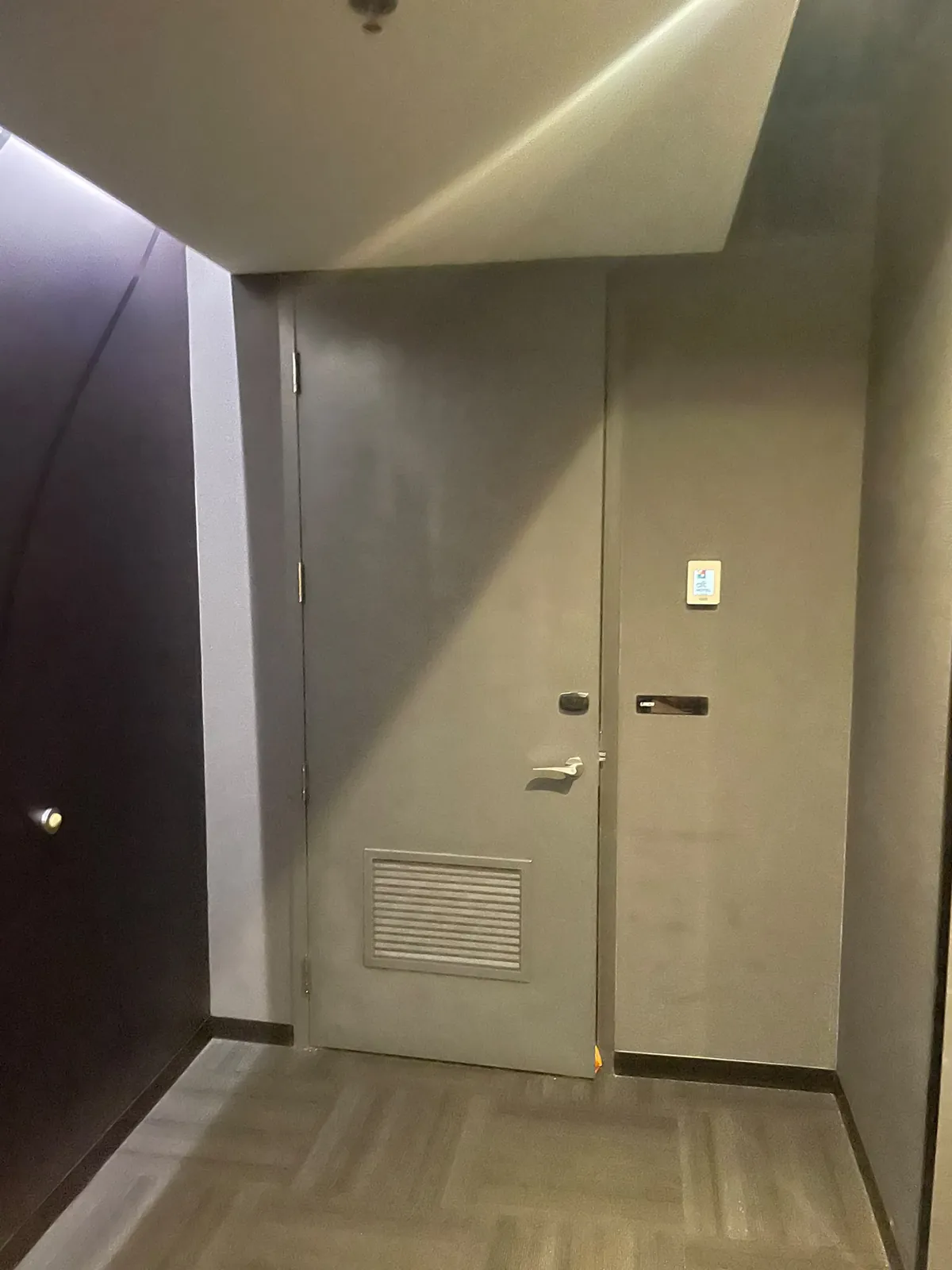 End of a guest corridor with a grey service door, a louvred vent and a cove-lit ceiling