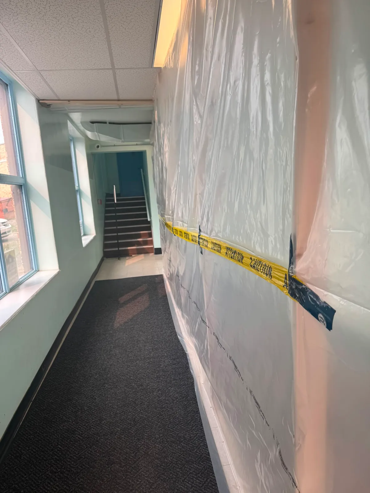 Corridor sealed off with floor-to-ceiling plastic sheeting and caution tape, leading to a stairwell