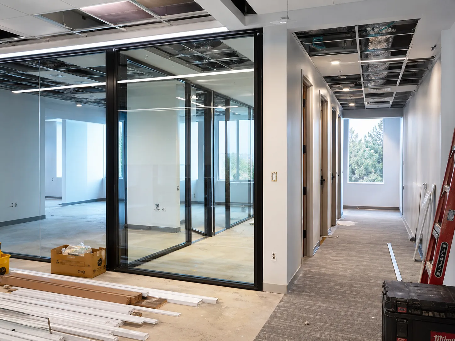Office floor mid-renovation with black-framed glass partitions, a new corridor of doors and an open ceiling grid
