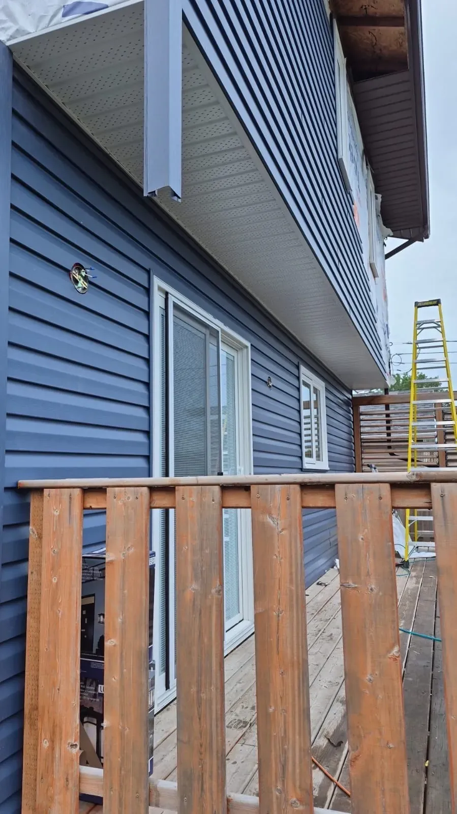 Blue vinyl siding and white soffit above a wood deck railing beside a sliding patio door