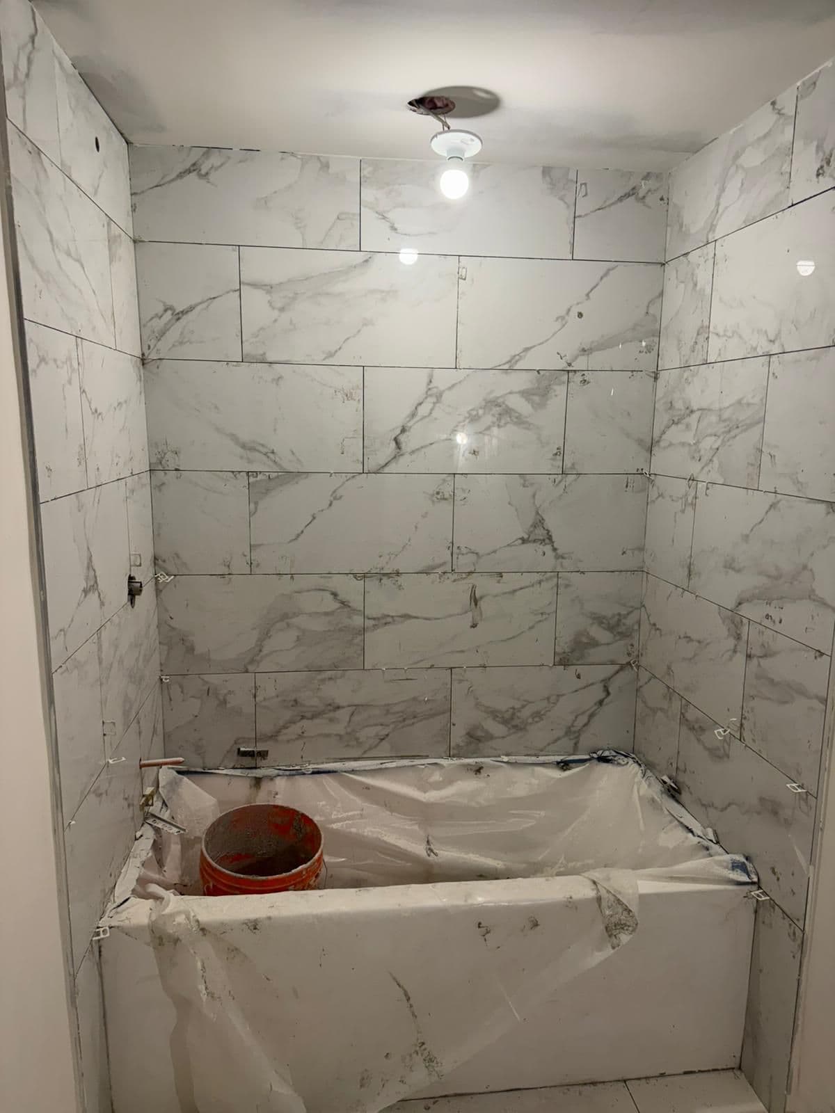 Large marble-look tiles set around a bathtub that is still wrapped in plastic, with tile spacers in the joints