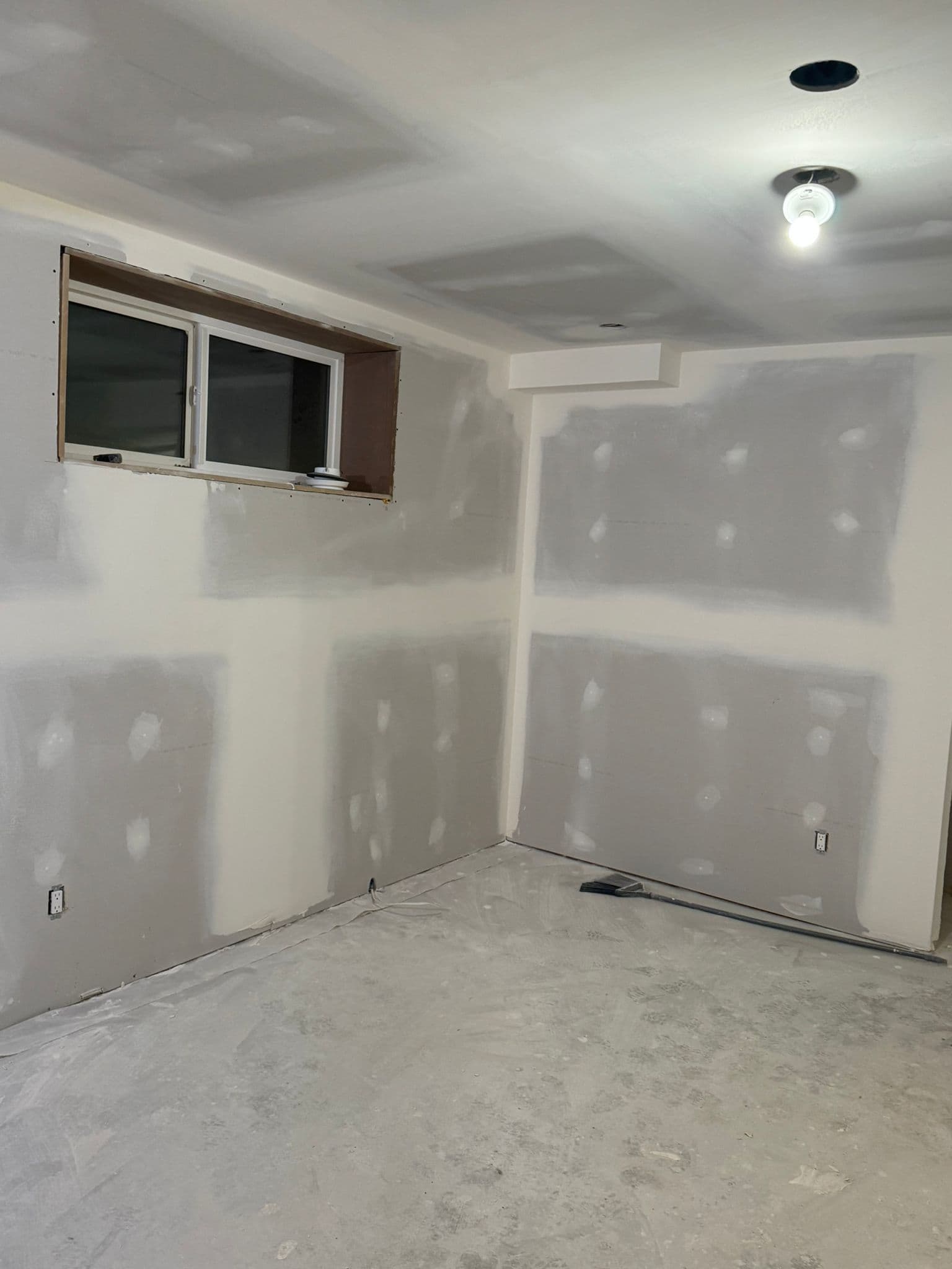 Basement bedroom with taped drywall, a window in a wood-lined opening, a ceiling light and a bare concrete floor