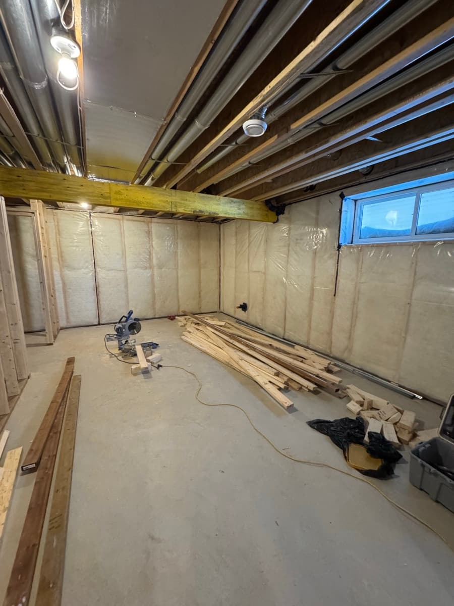 Unfinished basement with insulated foundation walls under clear poly, a wood beam, open joists, ducts and stacked lumber