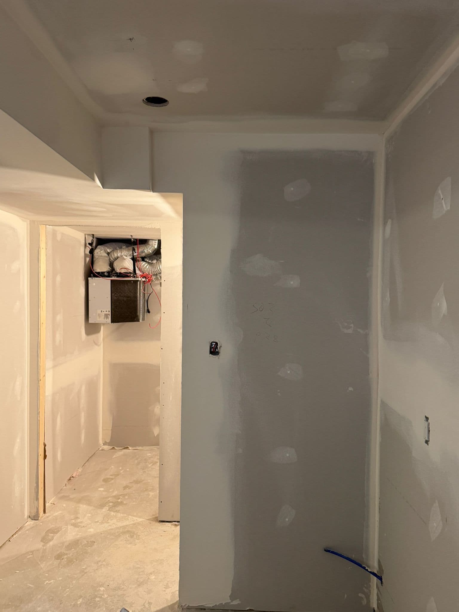Drywalled hallway beside a small mechanical room with a wall-mounted ventilation unit and insulated ducts