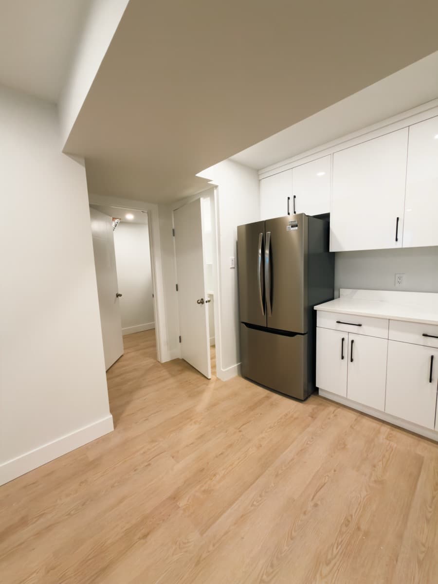 Finished basement hallway opening to a white kitchen with a stainless fridge and light plank flooring
