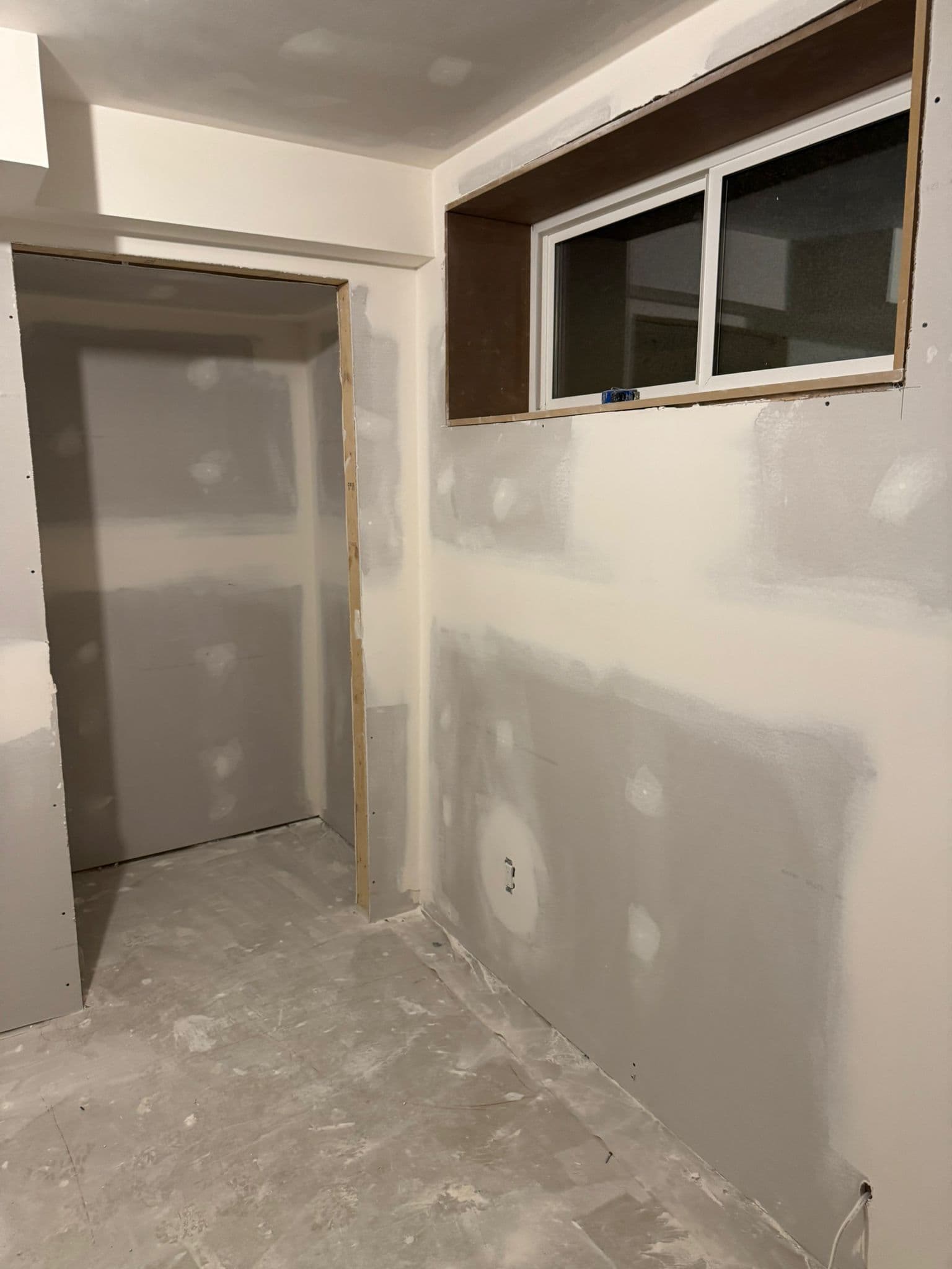 Basement room with taped drywall, a framed window opening, a doorway and a dusty concrete floor