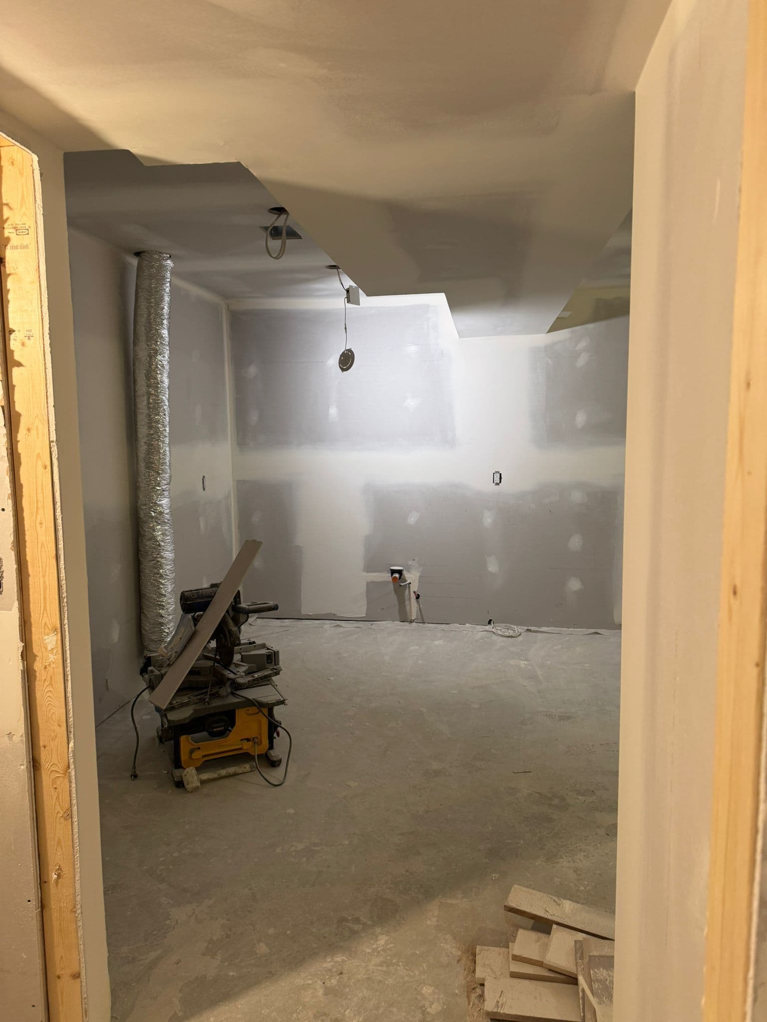 View through a door frame into a drywalled basement room with a flexible duct, capped water lines and a saw