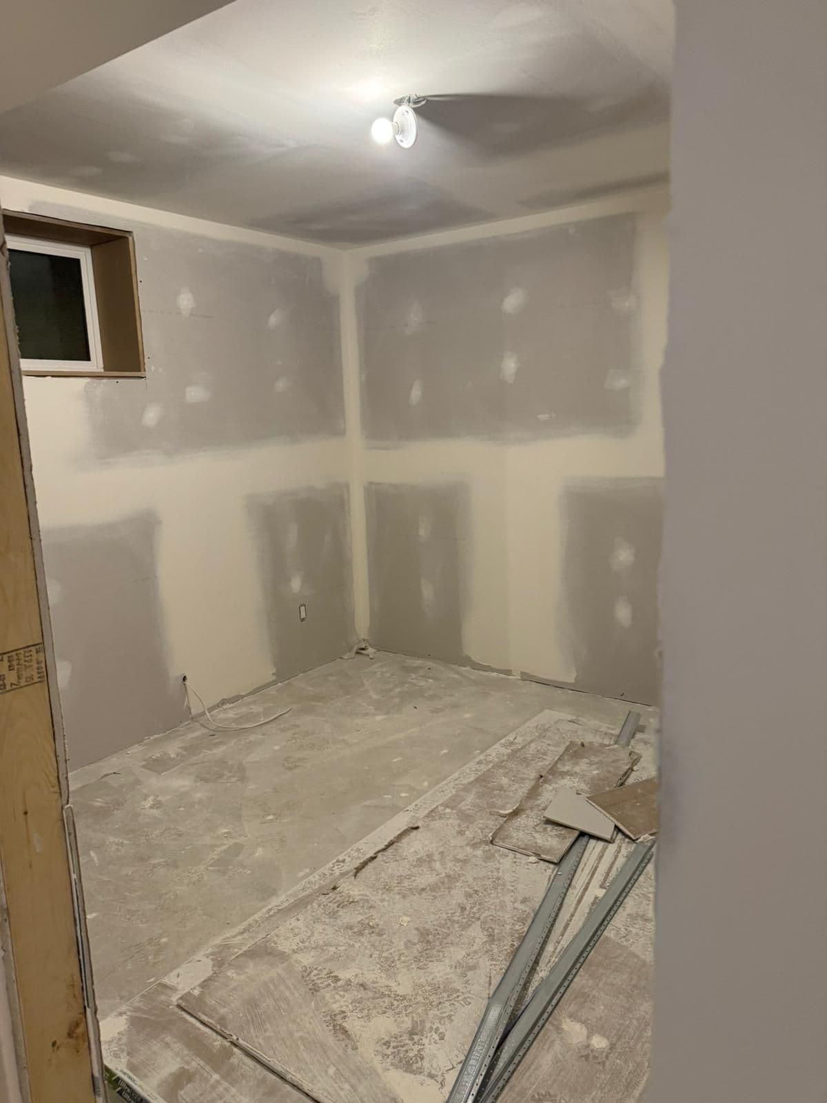 Small basement room with taped drywall on every wall, a window near the ceiling and a dusty concrete floor