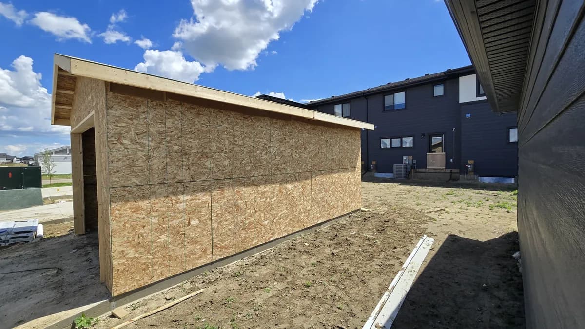 Side wall of the same garage in OSB sheathing on its foundation, with the backs of new townhouses behind it