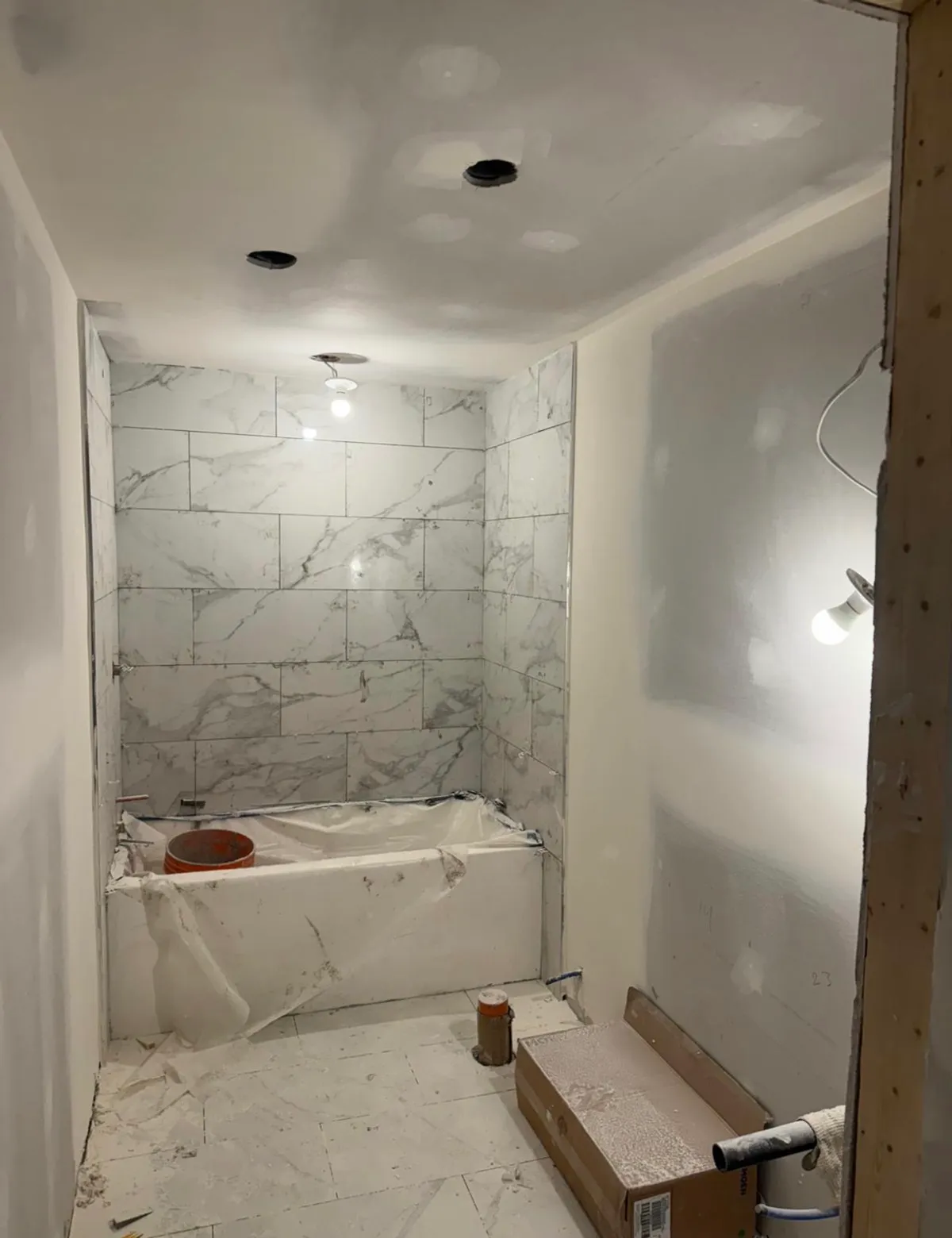 Bathroom under construction with marble-look wall tile set around a covered tub, taped drywall and floor tile laid