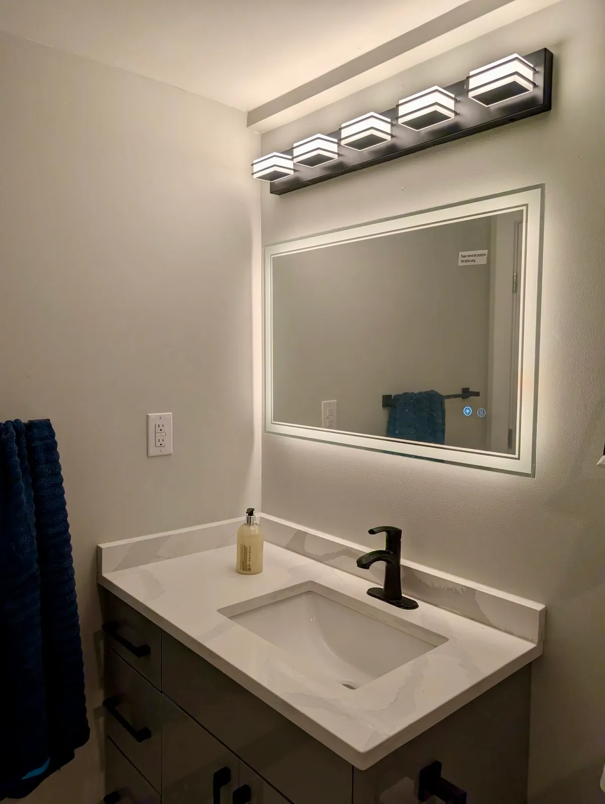Bathroom vanity with a white top and black faucet under a backlit mirror and a row of vanity lights