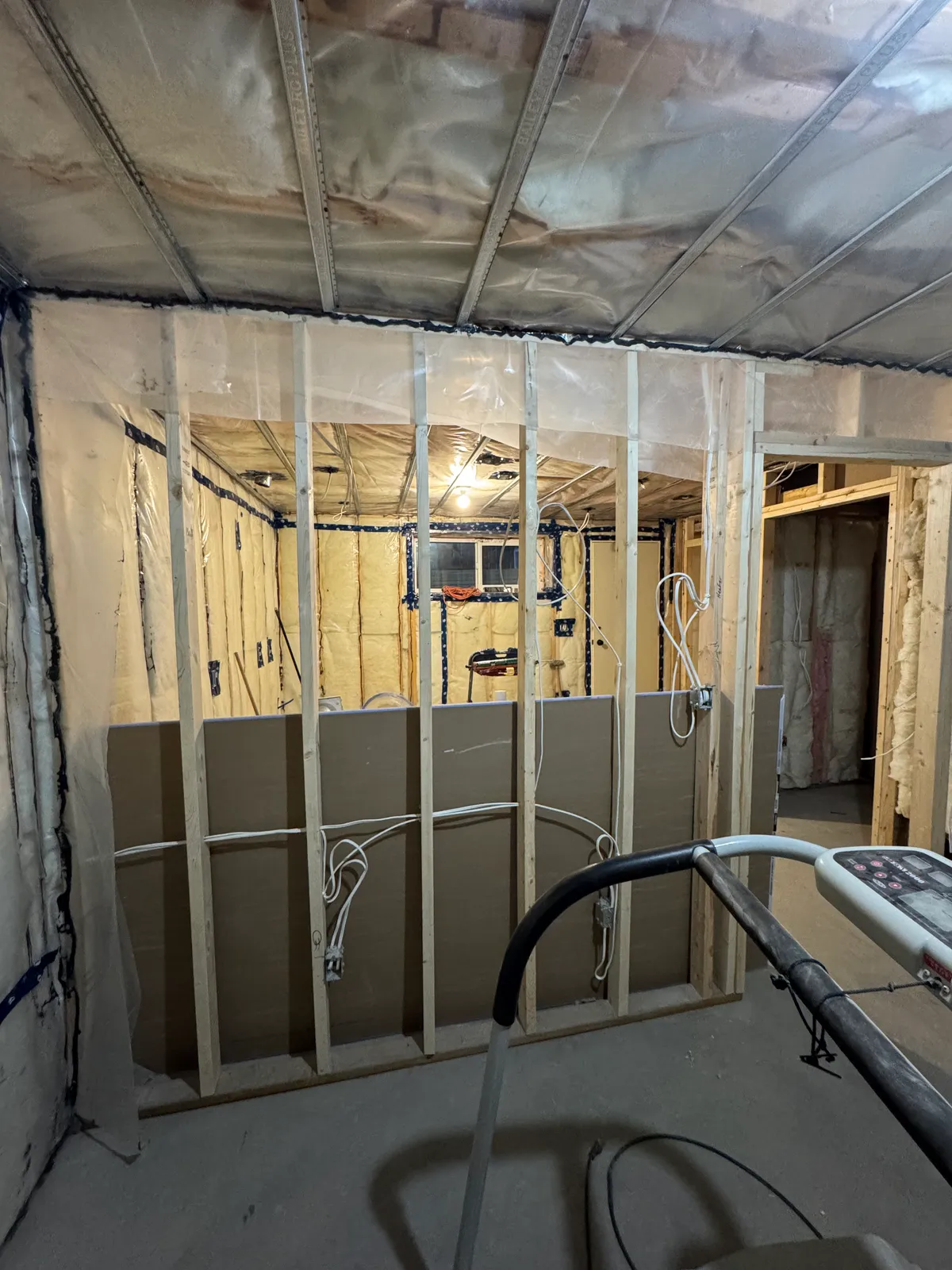Basement under construction with insulated walls behind poly, new stud framing, loose wiring and drywall started on the lower wall
