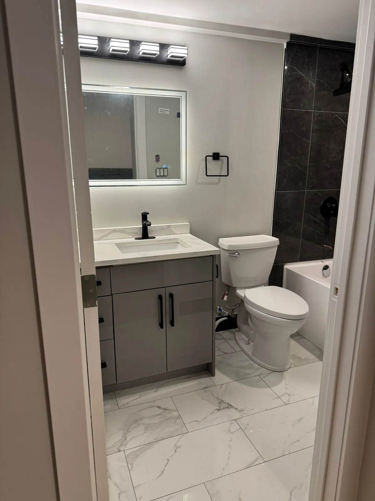 Suite bathroom with a grey vanity, a backlit mirror, a toilet and dark tile around the tub