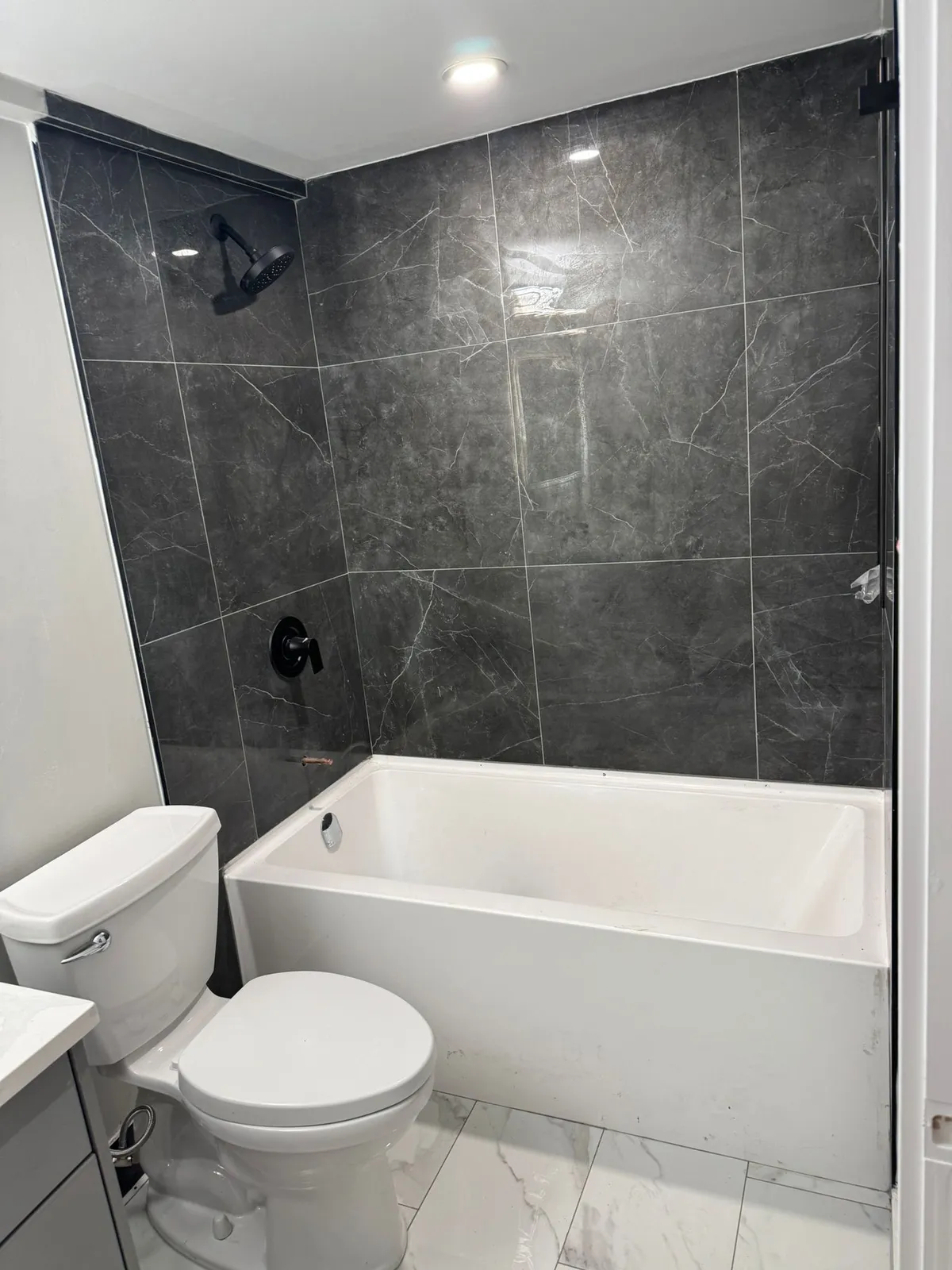 Basement bathroom with dark marble-look wall tile around a white tub, a toilet and white floor tile