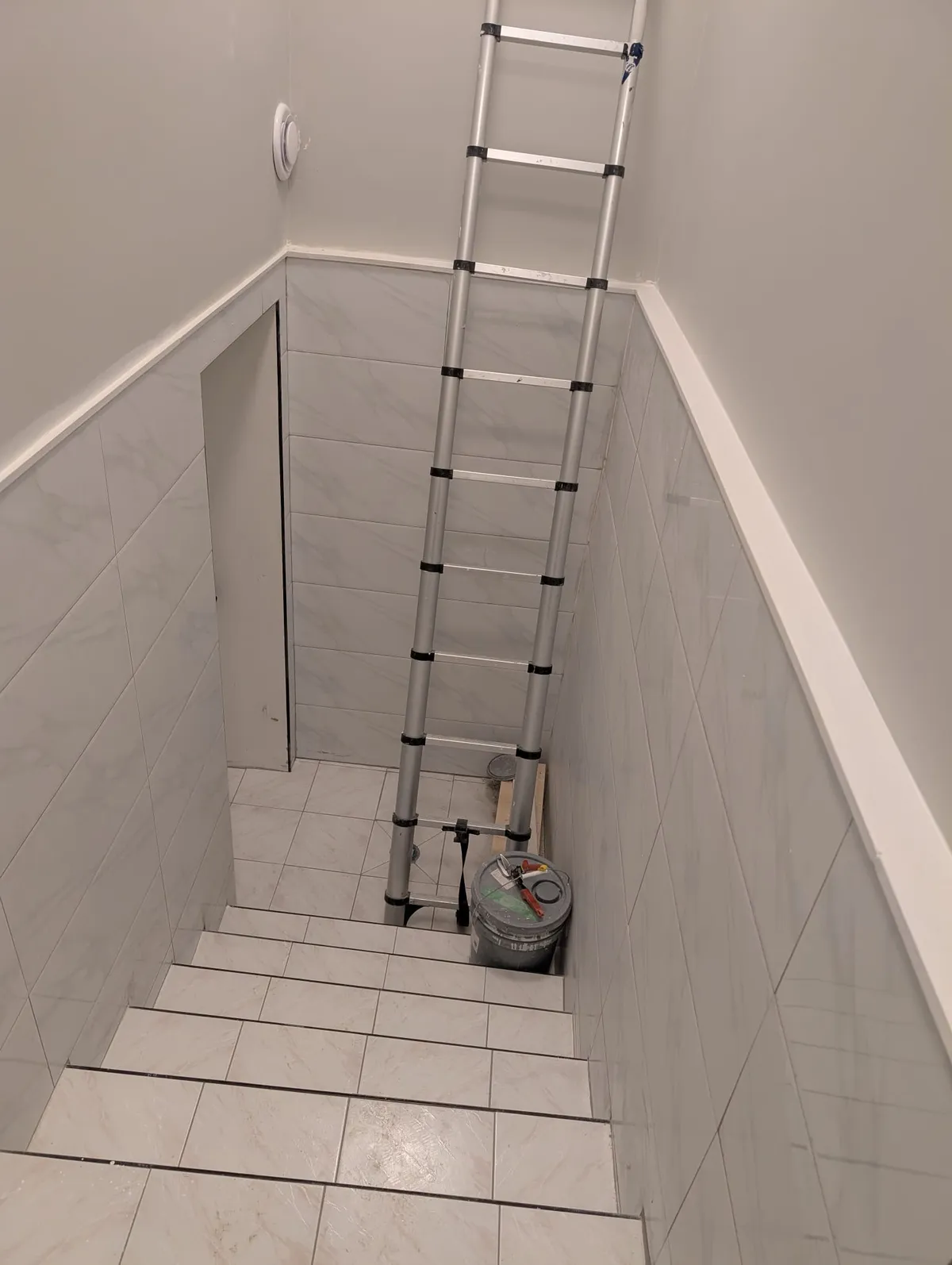 Stairwell with white tile on the steps and lower walls, and a ladder and paint pail at the bottom