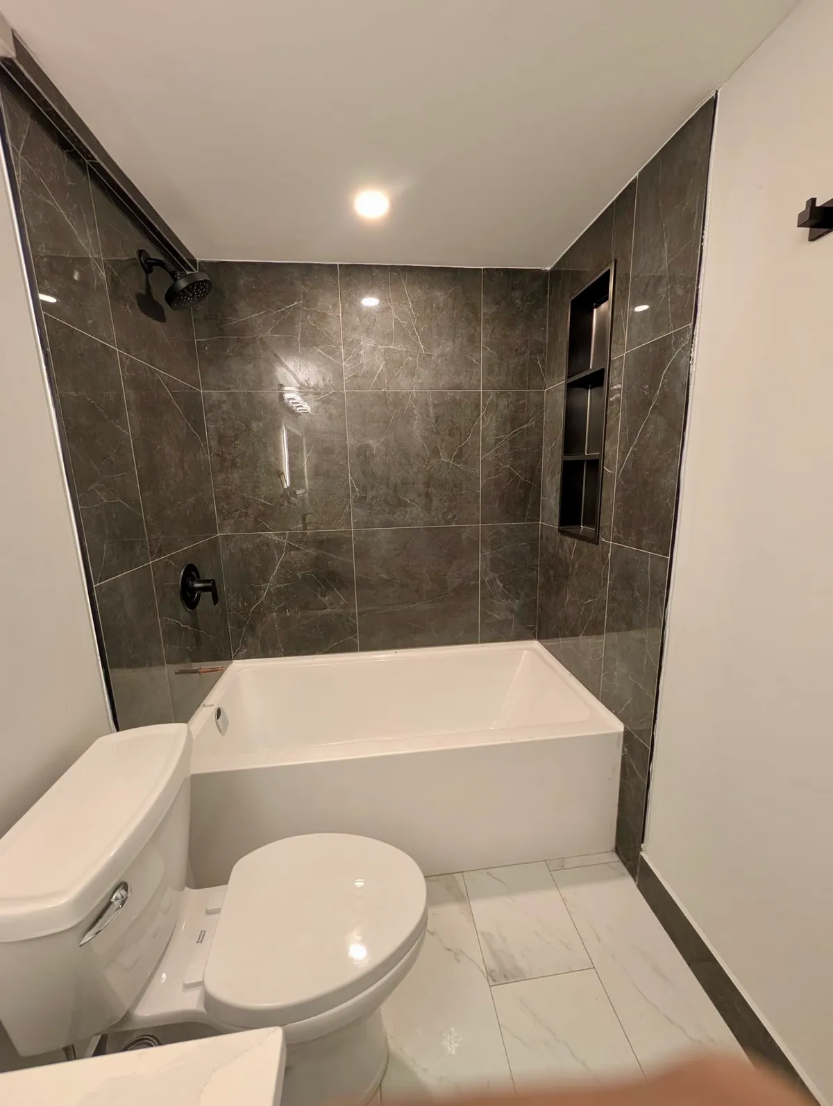 Tub with dark grey stone-look wall tile and a recessed niche, beside a white toilet and marble-look floor tile