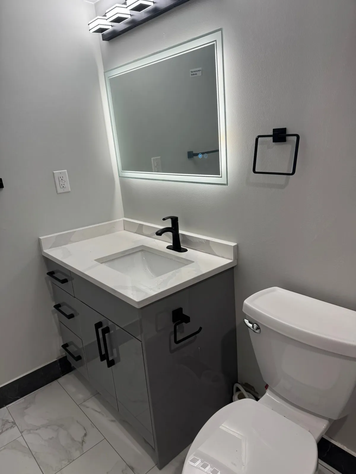 Grey bathroom vanity with a white top, a black faucet and a backlit mirror above it