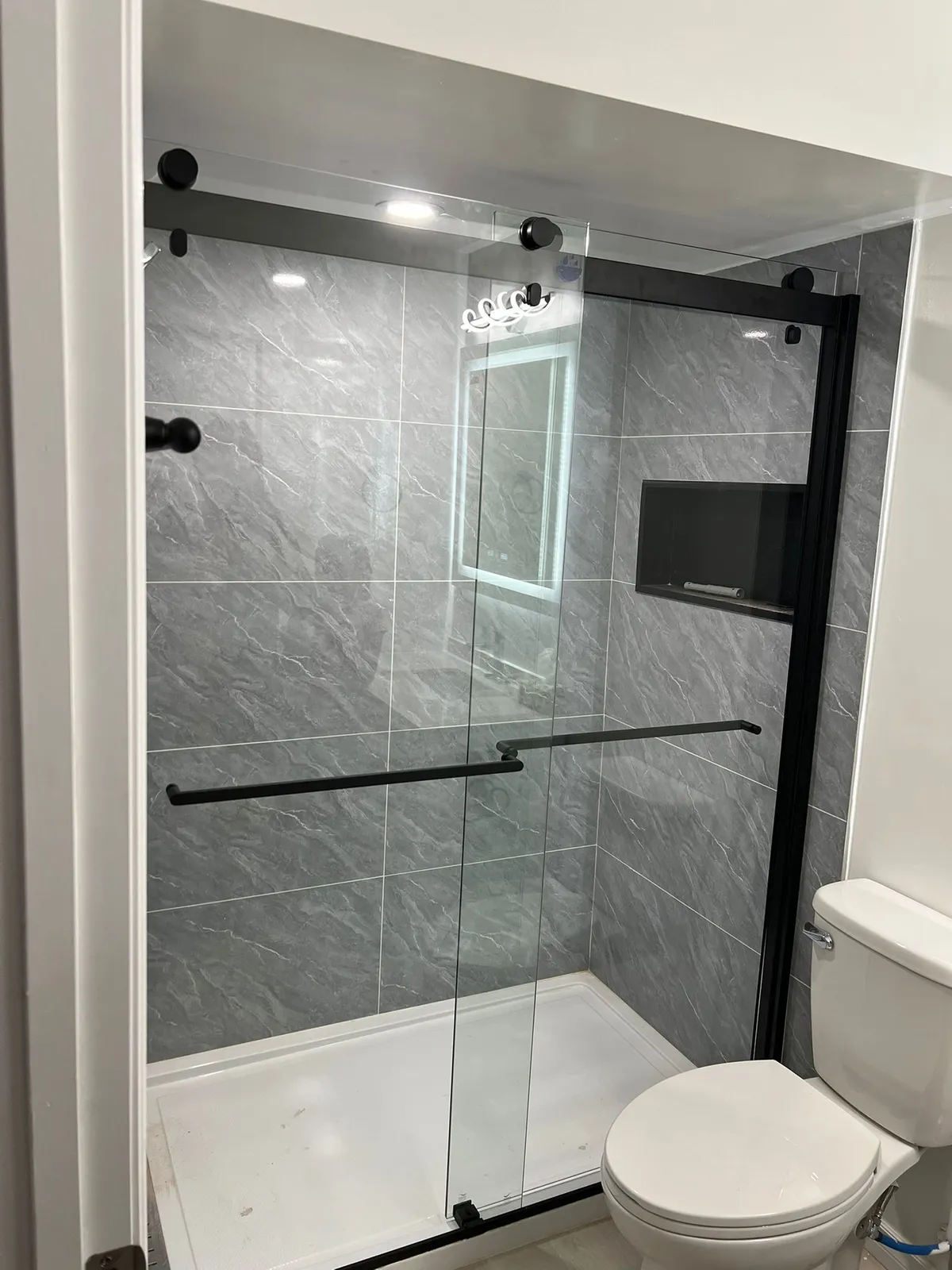 Straight-on view of the shower, with large grey marble-look tiles, a black wall niche and black towel bars on the glass