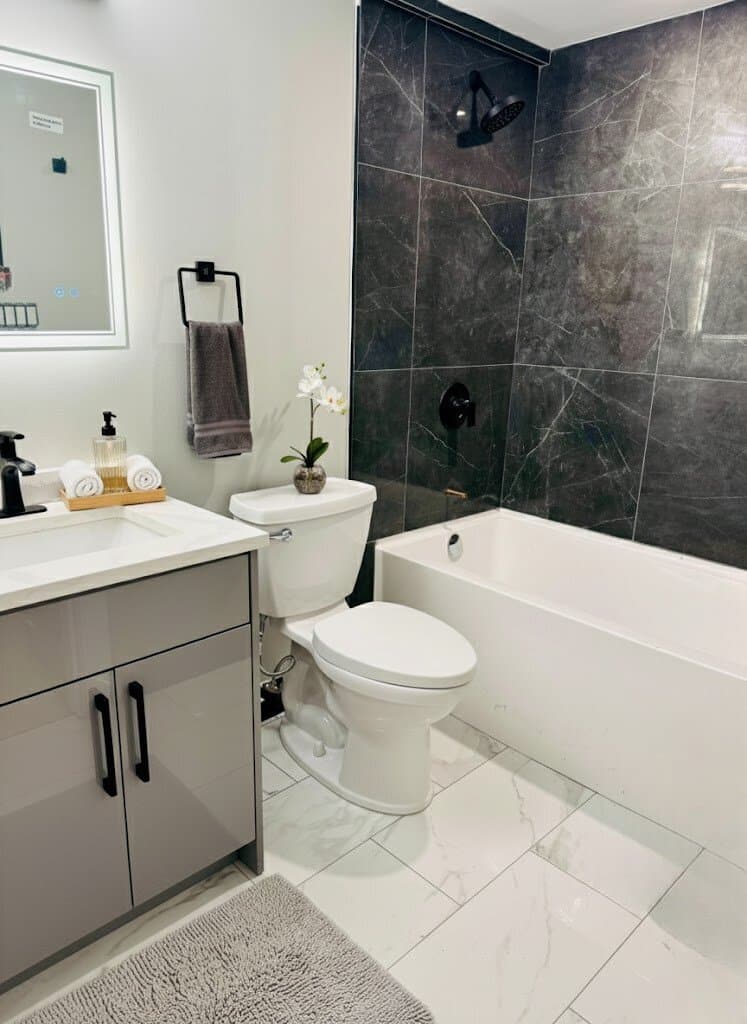Bathroom with a grey gloss vanity, lit mirror, white alcove tub and dark marble-look wall tile with black fixtures