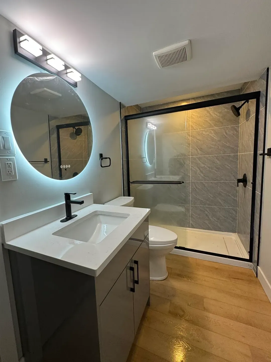 Renovated bathroom with a backlit round mirror, a white quartz vanity and a black-framed glass shower door